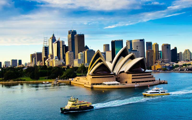 "An aerial view of the Sydney Opera House and the surrounding cityscape, with the iconic white sails of the Opera House visible in the foreground. The image also features the glistening waters of the Sydney Harbour, with boats sailing along the river."