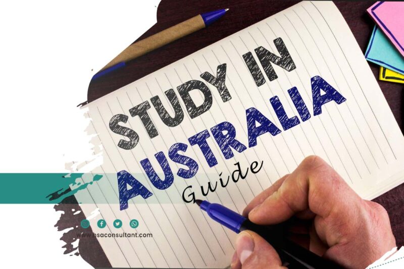notebook lies down horizontally on the table. A hand is holding a pen & "Study in Australia Guide" is written on notebook